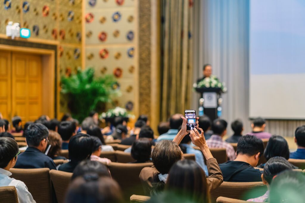 Back side of audience taking photo by cellphone the Speaker with podium and presentation